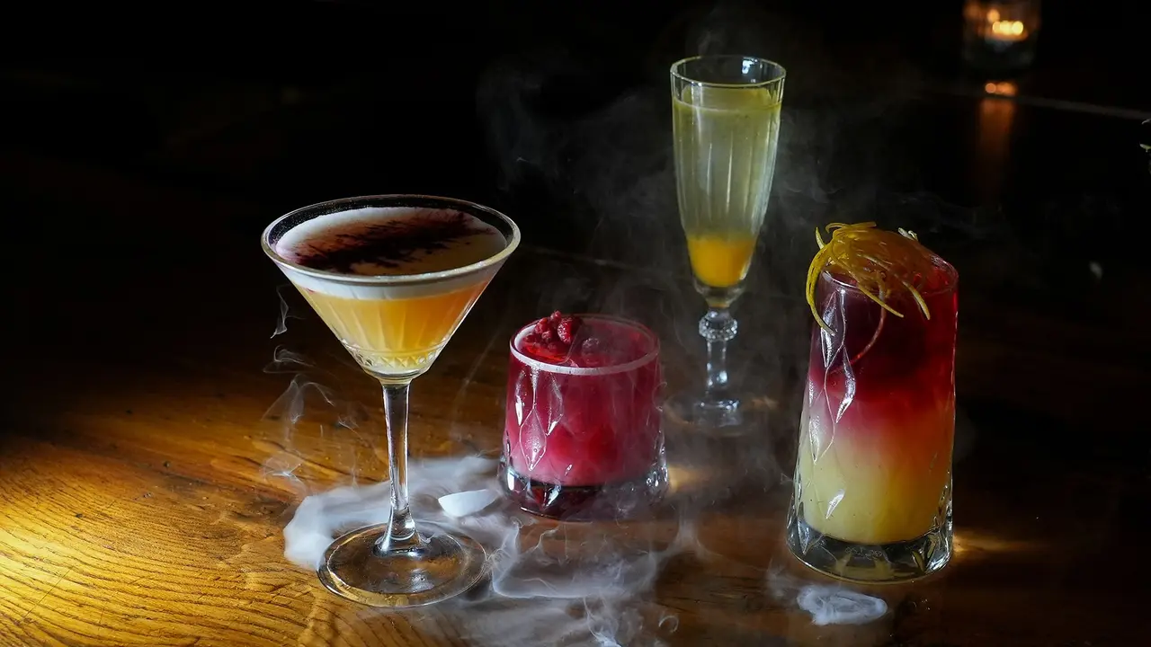 Four signature cocktails on a wooden bar with swirling smoke